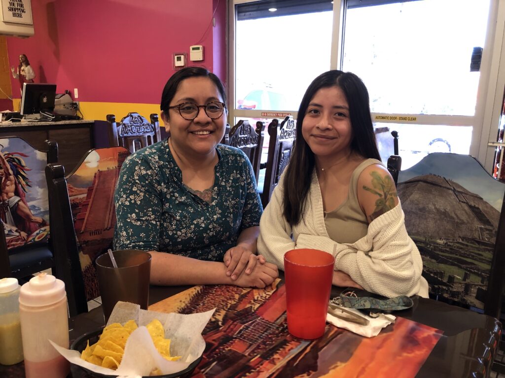 Two women, one in blue, one in white, sit at a wooden table with tortilla chips and a red plastic water glass.