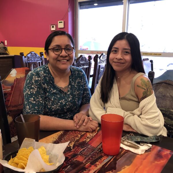 Two women, one in blue, one in white, sit at a wooden table with tortilla chips and a red plastic water glass.