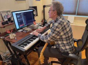 In this photo, Songs of Love Foundation's John Beltzer sits at an electronic piano keyboard and is typing on a computer keyboard that's on top of it. He's looking at a computer monitor that's on a desk behind the piano keyboard. A guitar rests in his lap.