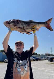 A man wearing a hat and glasses holds a fish above his head.