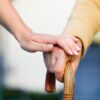 A person puts their hand over an older person's hand that's holding a cane.