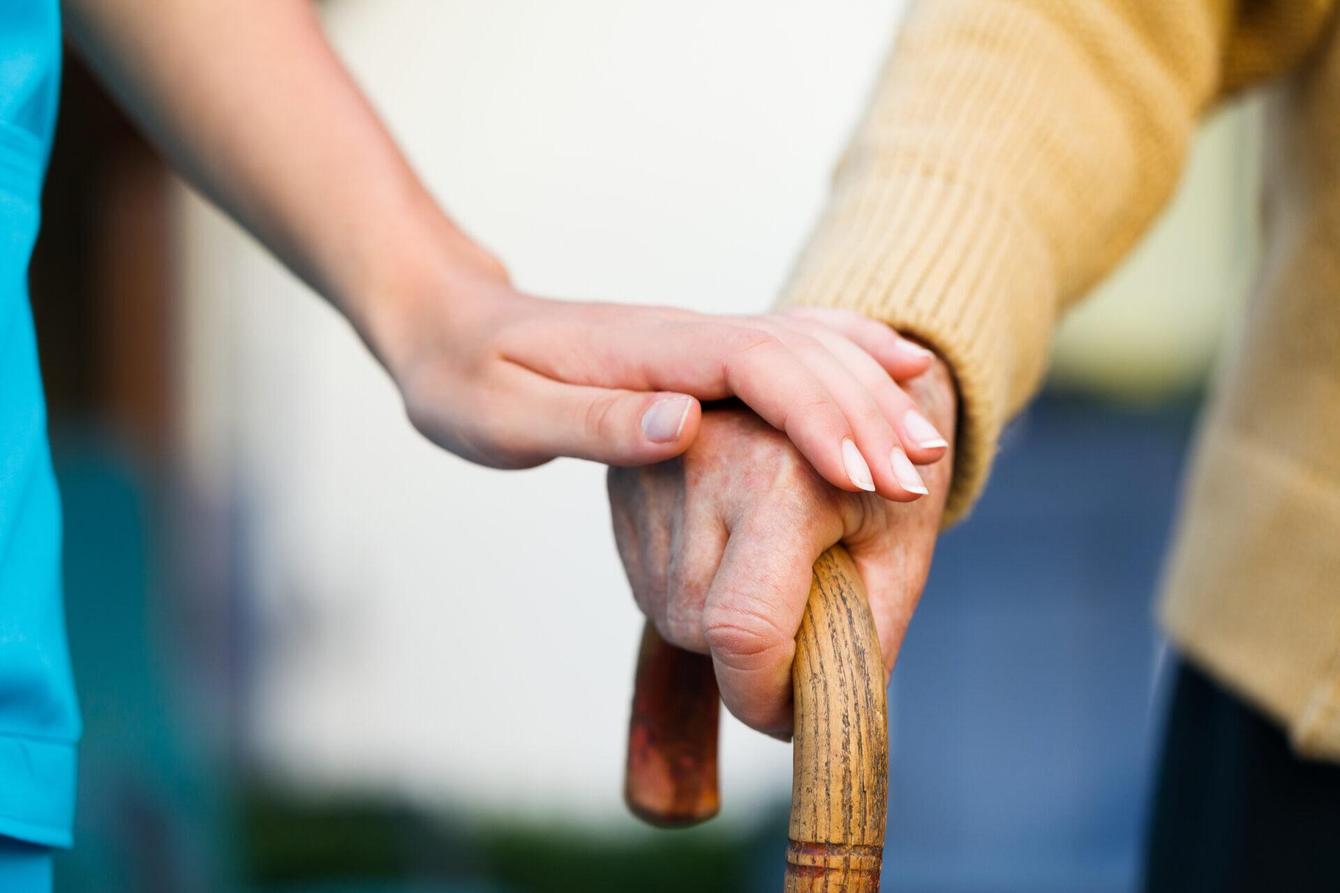 A person puts their hand over an older person's hand that's holding a cane.