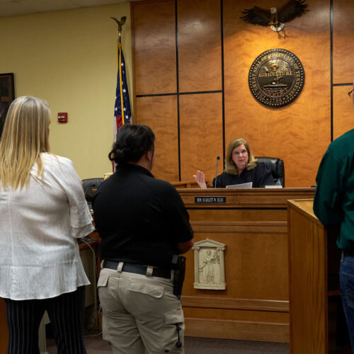 A courtroom scene in which three women and one man stand facing a judge. The judge is gesturing with her hand as she speaks to the group. The three women are on one side of the room, and the man is on the other.