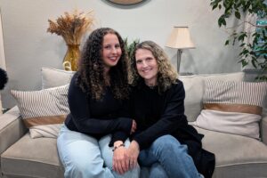 A teenager with curly, brown hair sits next to her mother who has shoulder-length, curly blonde hair, on a couch. They are both wearing black sweaters with blue jeans.