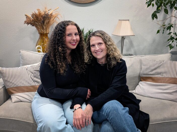 A teenager with curly, brown hair sits next to her mother who has shoulder-length, curly blonde hair, on a couch. They are both wearing black sweaters with blue jeans.