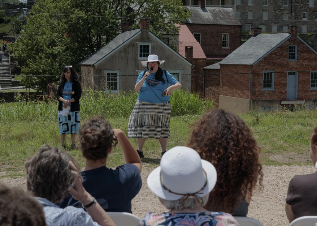 Former National Park Ranger Melissa Dalley, 49, seen here holding a microphone in front of a crowd, speaks during the America 433+ teach-in at Harpers Ferry National Historic Park on June 19, 2026.