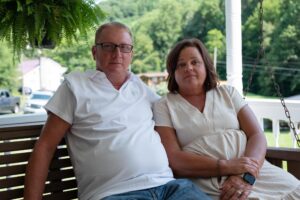 Tim Balthis, 57, and his wife, Fran, sit outdoors at their home in Pound, Virginia. He's wearing a white, short-sleeved shirt and glasses; she's wearing a light-colored dress.