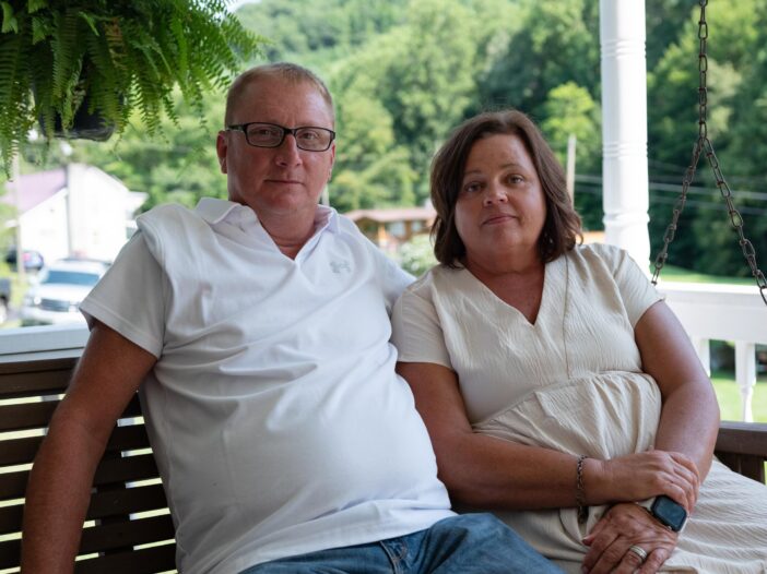 Tim Balthis, 57, and his wife, Fran, sit outdoors at their home in Pound, Virginia. He's wearing a white, short-sleeved shirt and glasses; she's wearing a light-colored dress.