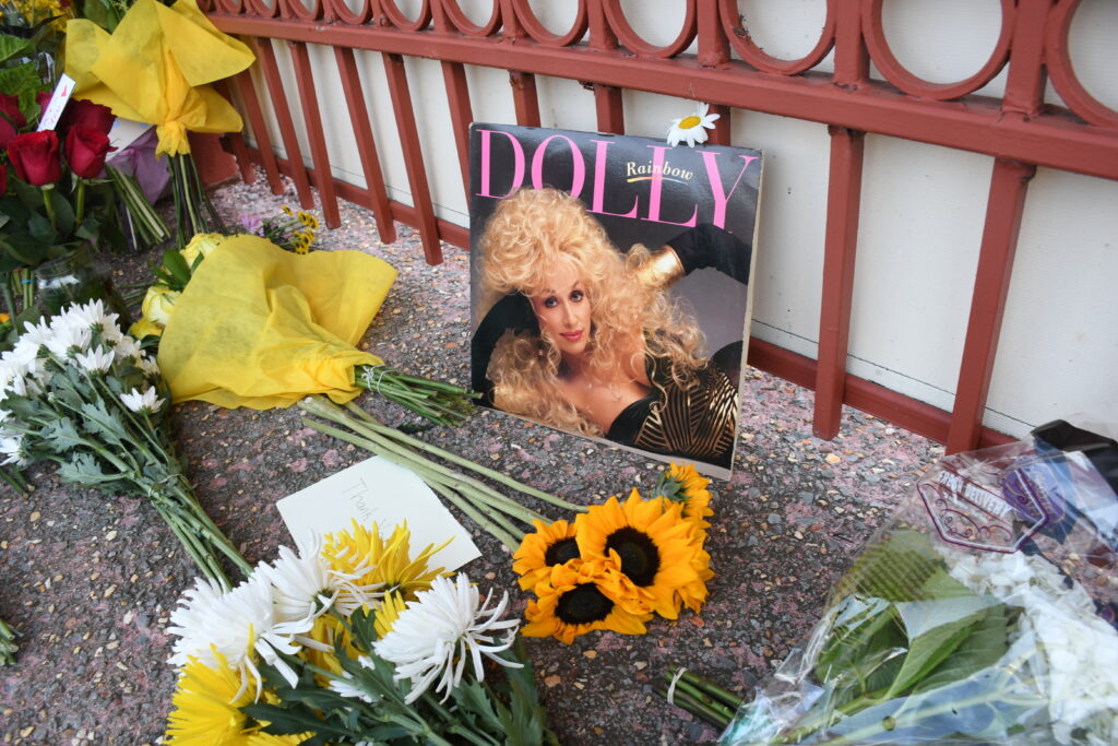 Photo of a young Dolly Parton is propped against a gate and surrounded by flowers.