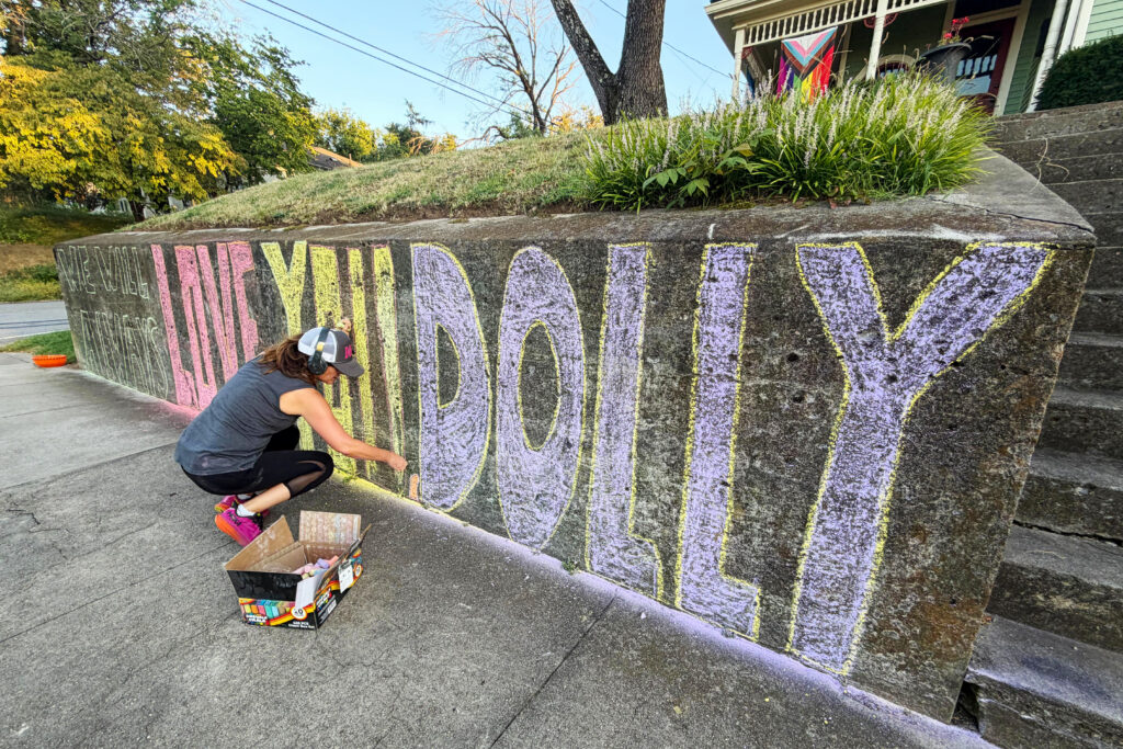 A woman with headphones and a ballcap on as she spells 'We will always love you Dolly' in large text on concrete with colorful chalk.