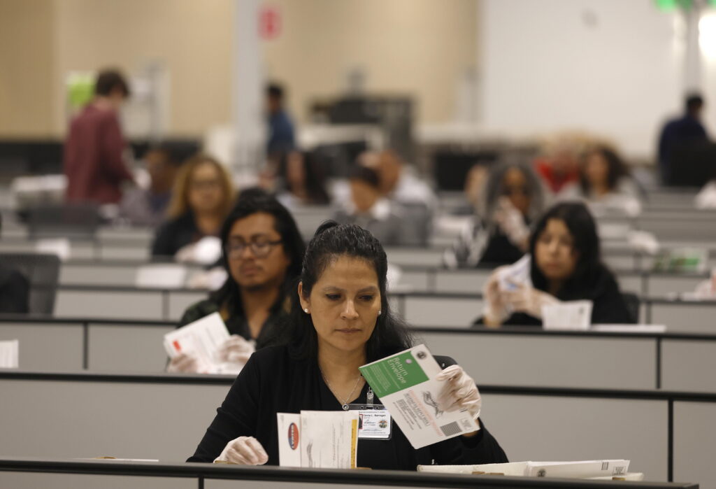 Election workers who are seated and wearing clear gloves process ballots for the California state primary.